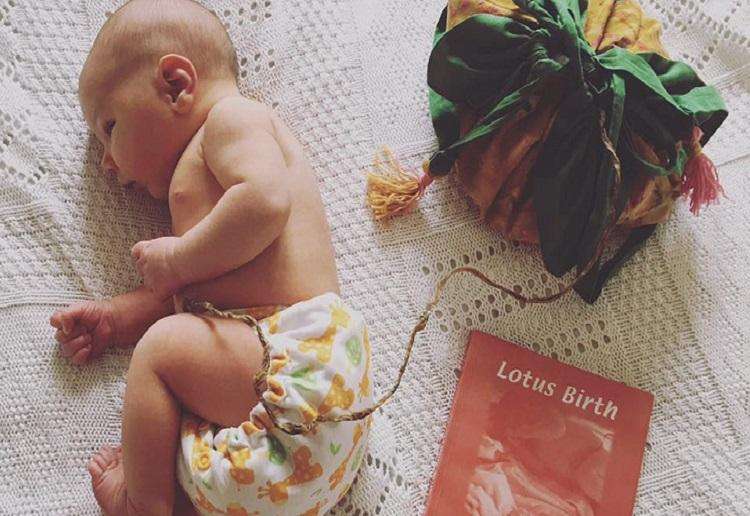 A baby lying on a white blanket beside a wrapped lotus birth bundle and a card reading Lotus Birth