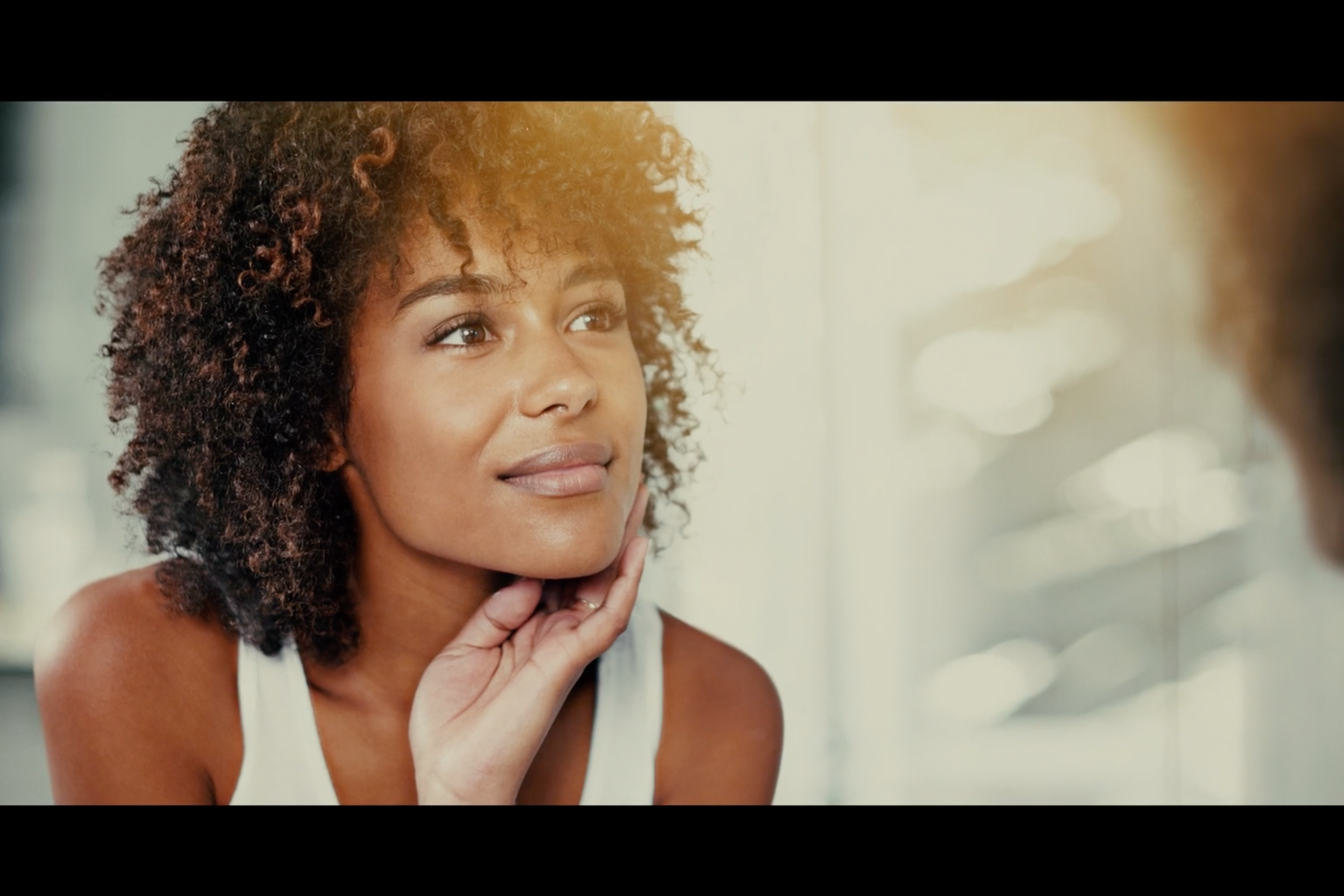 A woman with curly hair rests a hand at her jaw and looks at her own reflection in a mirror