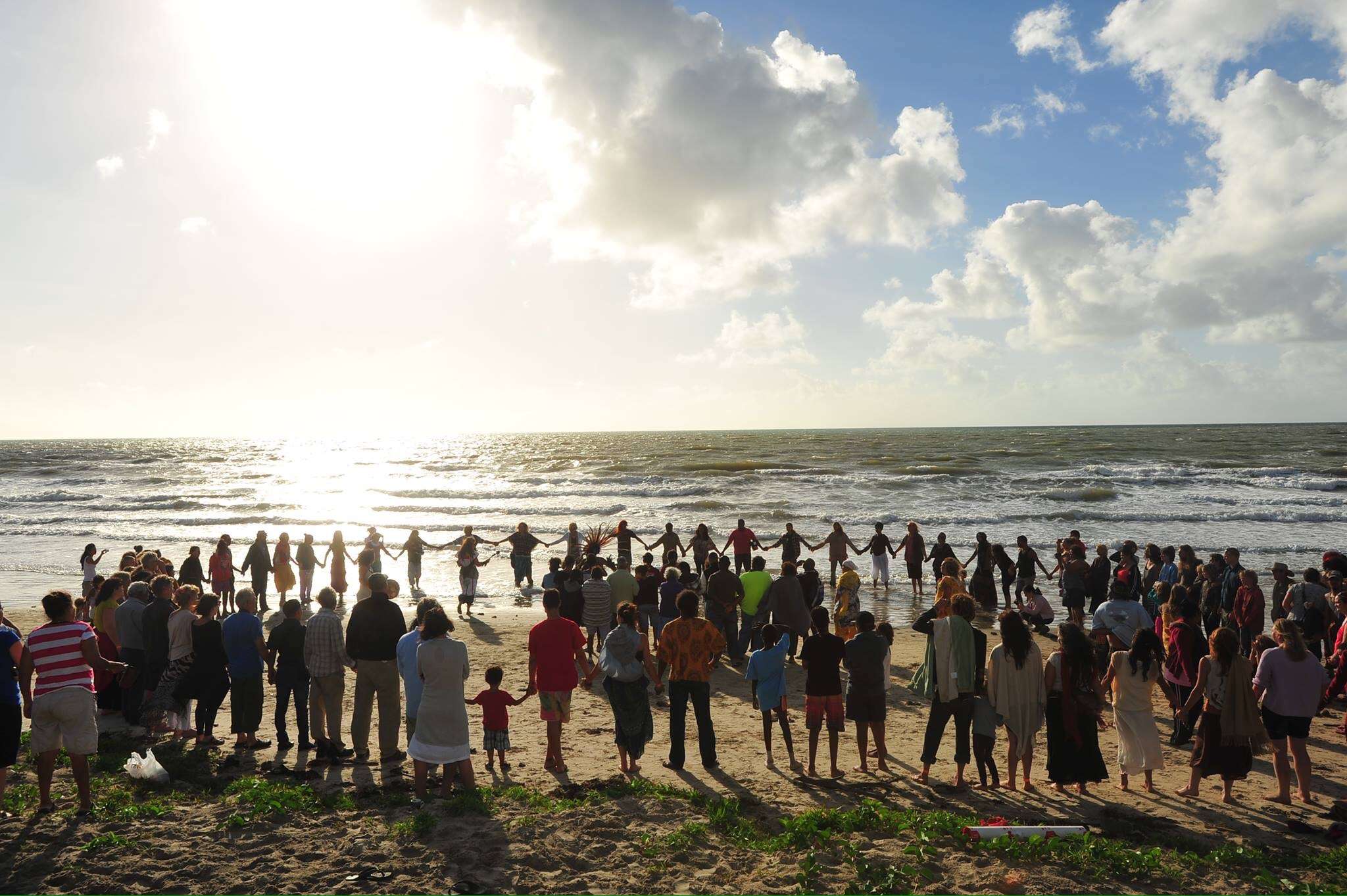 A large circle of people standing hand in hand on a wet beach at dawn, the sea and a bright horizon behind them.