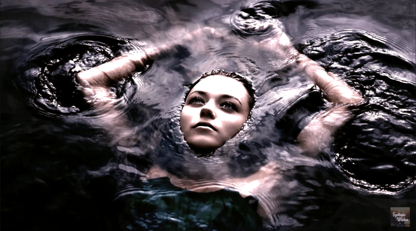A figure lies in dark water with arms lifted above the surface, face turned toward the light