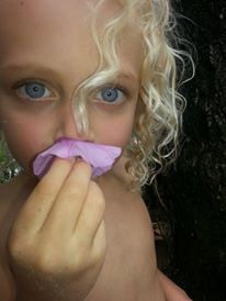 A fair-haired child holding a pink petal to their lips.